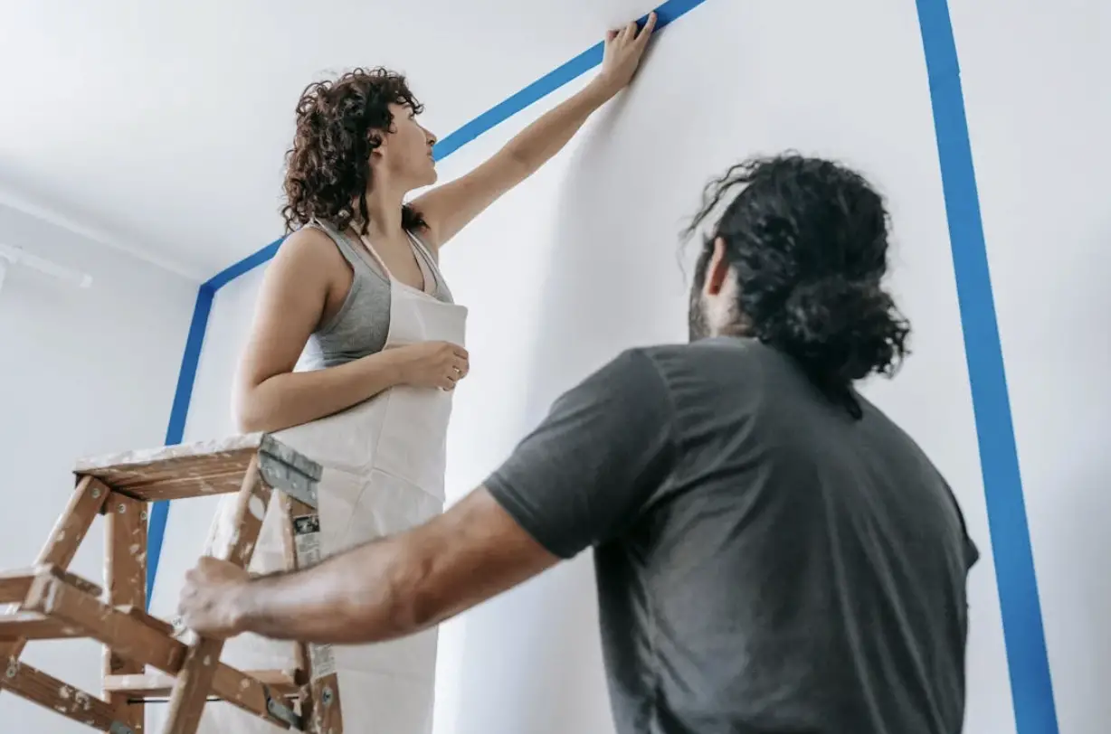 A girl standing on a ladder putting up paint tape. With a guy standing below looking at her.